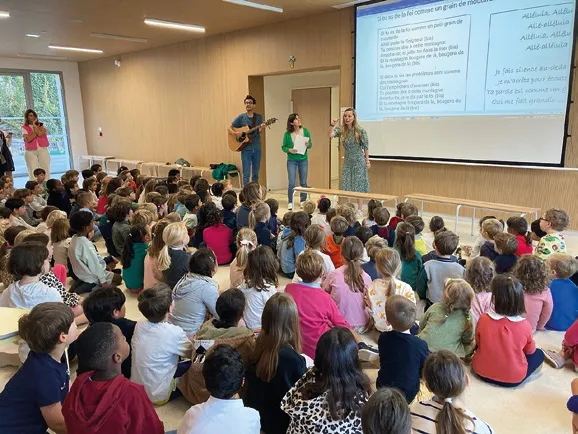Toute l'école réunie dans la grande salle, de dos, écoute trois adultes qui chantent à la guitare.
